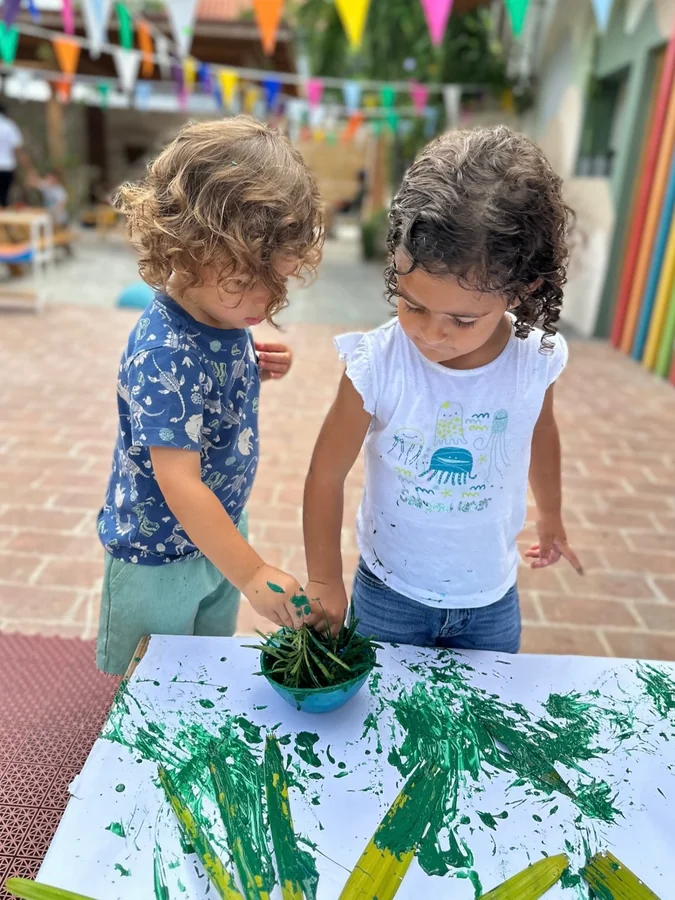 Dos niñas pintan con hojas sobre un papel grande en el patio