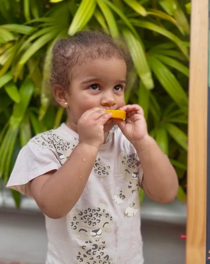 Una niña pequeña prueba una fruta al aire libre, rodeada de plantas